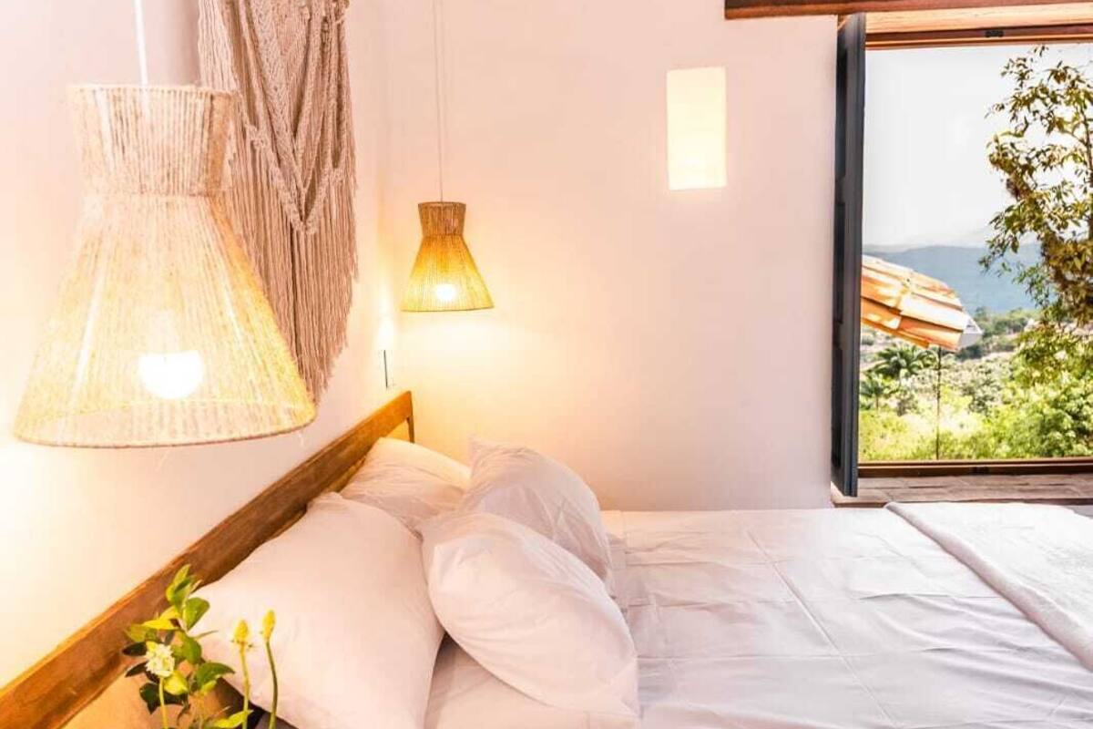 Una cama con sábanas blancas, una lámpara colgante de macramé y vistas a la vegetación por una ventana.
