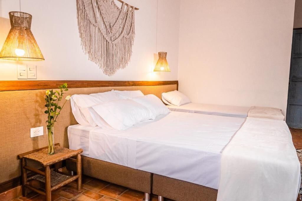 Hotellihuone, jossa on sänky, yöpöytä, lamppu, seinälle ripustettu makrame-koriste ja pieni kasvi.