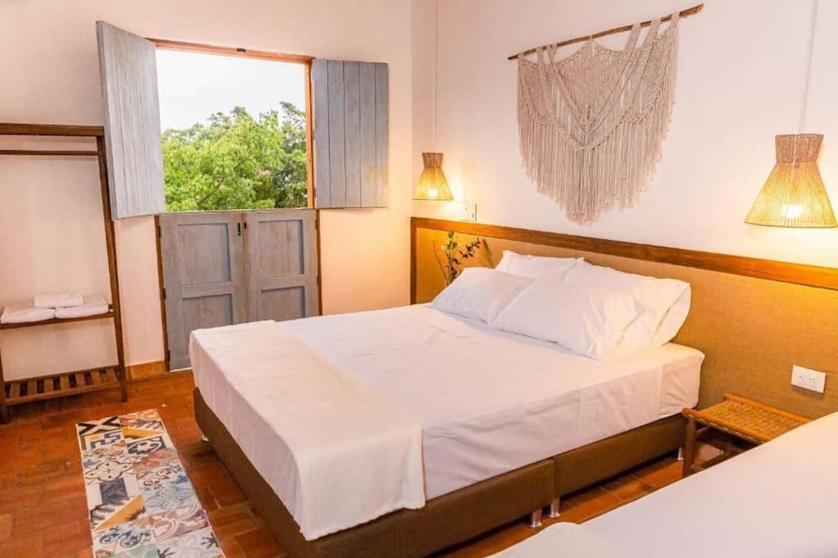 Un dormitorio con cama, una ventana con contraventanas abiertas, un adorno de macramé en la pared y una lámpara de noche.
