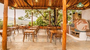 An outdoor dining area with wooden tables and chairs, a brick oven, and a view of the landscape. - CASA GUATI (Barichara)