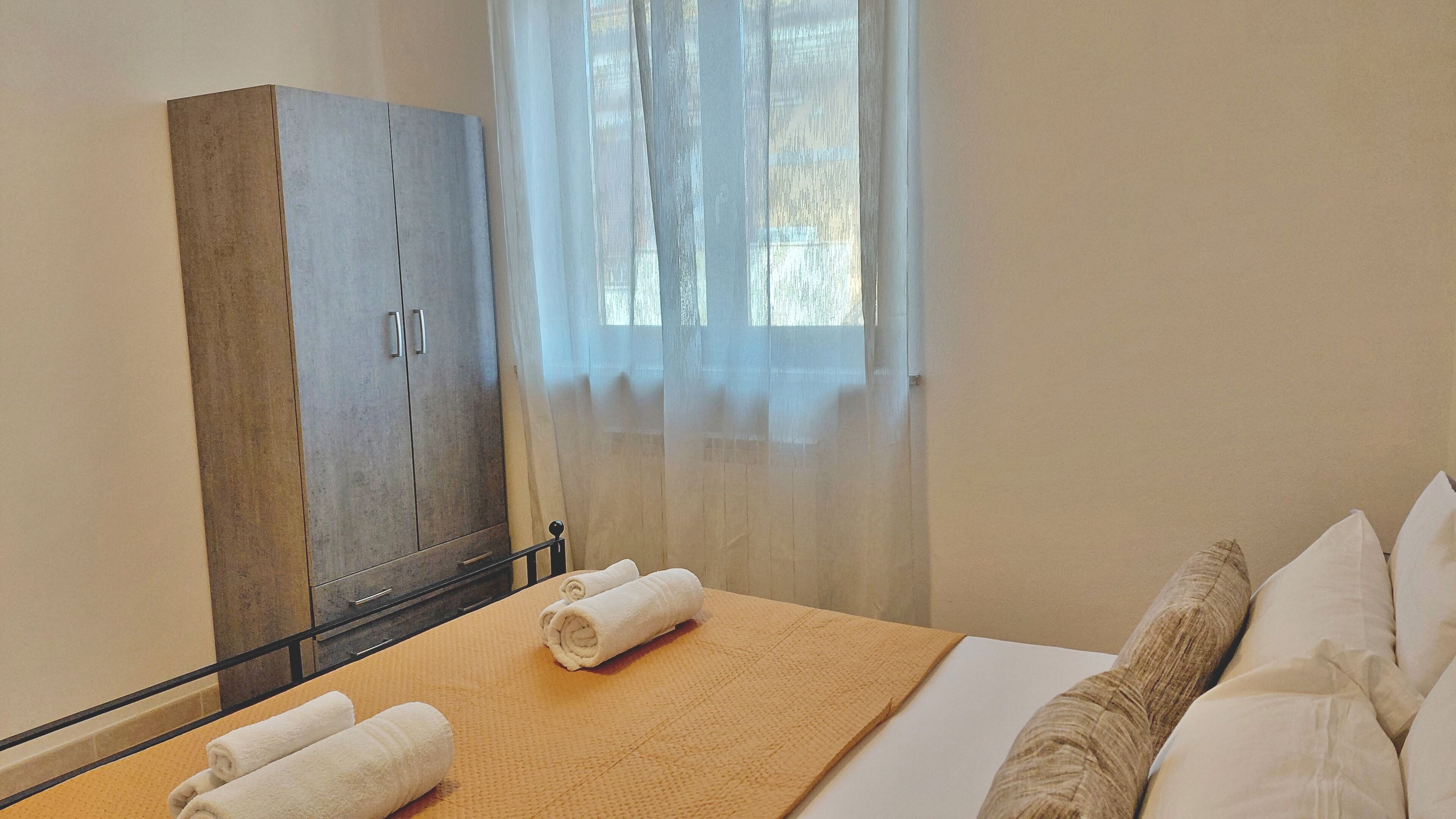 Une chambre d’hôtel avec un lit, une armoire en bois, une fenêtre avec des rideaux légers et deux serviettes enroulées posées sur un tapis.