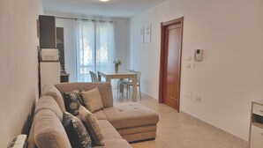 Ein Wohnzimmer mit einem beigefarbenen Sofa, einem Esstisch mit Stühlen, einer Holztür und einer Küchenzeile mit einem Kühlschrank.