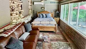 Un dormitorio rústico con pared de piedra, cama de madera, sofá de cuero y una ventana grande con vistas al exterior.