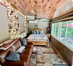 A rustic bedroom with a stone wall, a wooden bed, a leather sofa, and a large window offering a view of the outdoors. - Sleeps 14 on the River! Free Kayaks -by Whitewater (Canyon Lake)