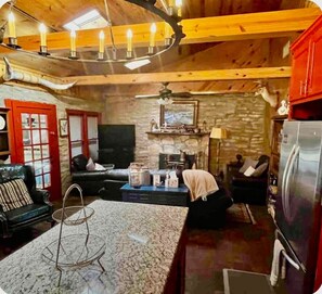 A cozy living room with a fireplace, a stone wall, and a modern kitchen with a refrigerator. - Sleeps 14 on the River! Free Kayaks -by Whitewater (Canyon Lake)