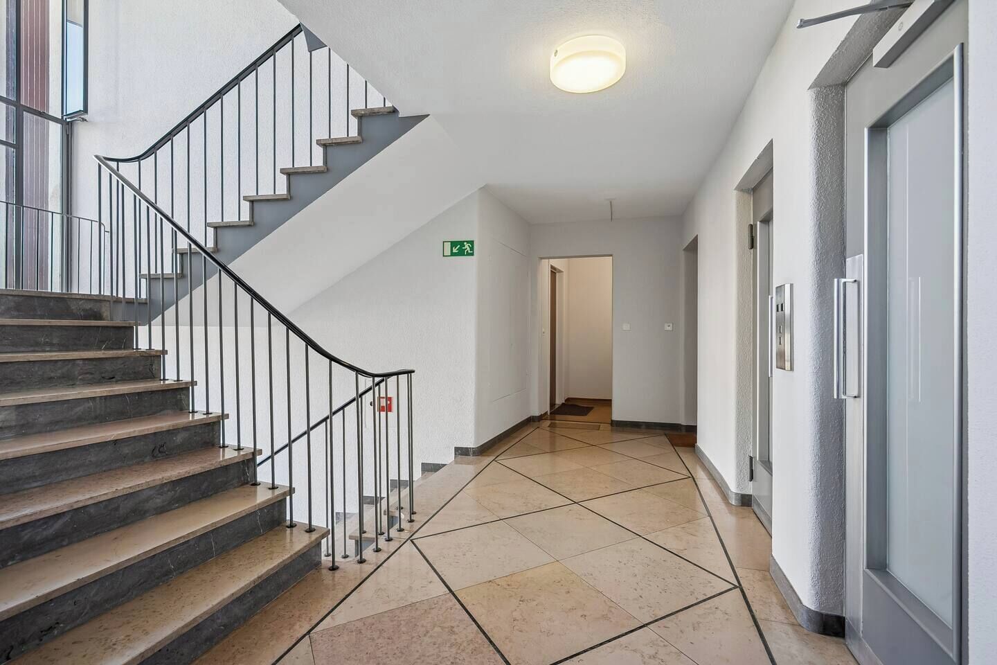 Een schone, moderne hal met een trap naar een bovenverdieping. Een groen bord met de tekst ‘uitgang bij nood’ is zichtbaar aan de muur.