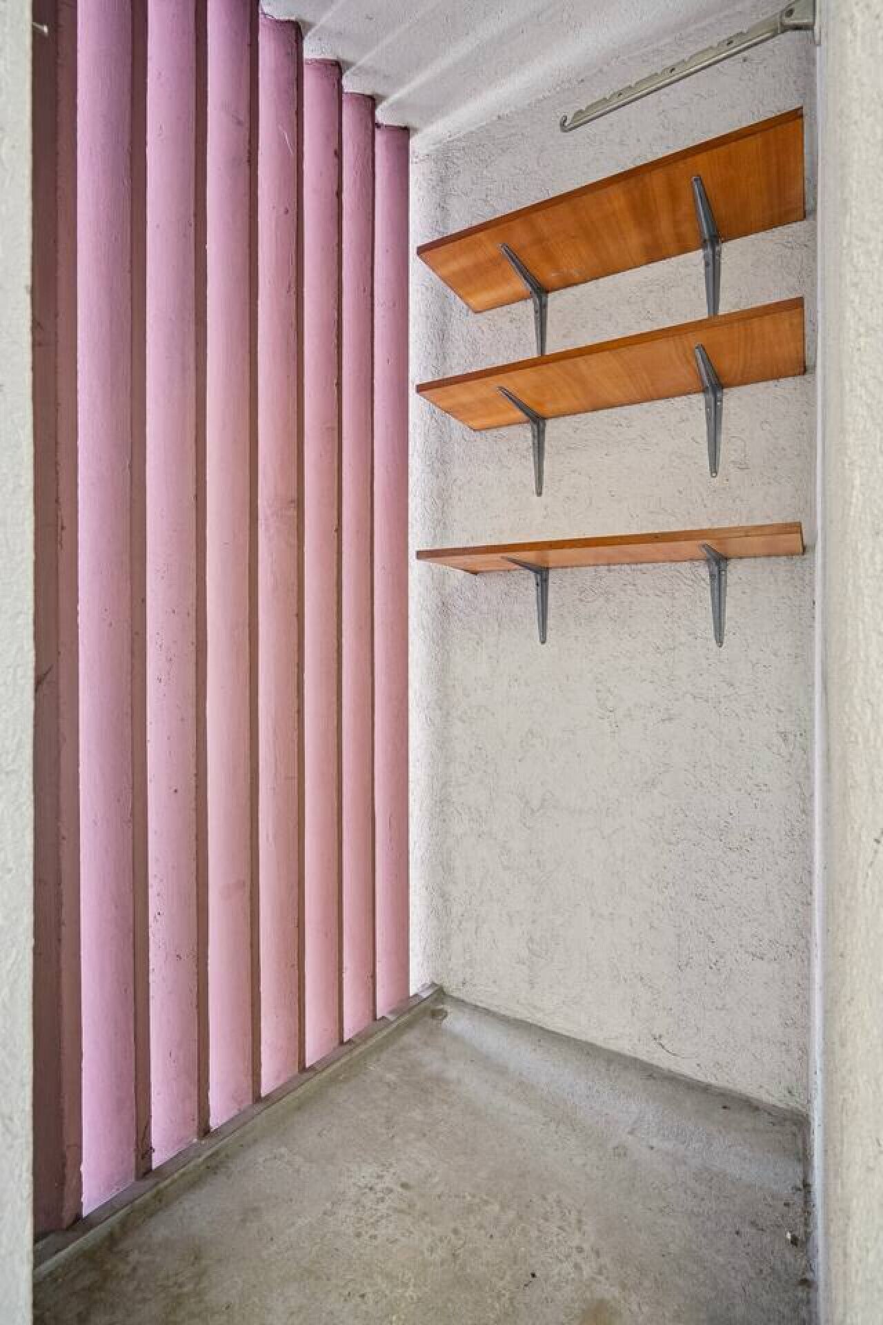Une pièce avec des stores verticaux roses, un sol en béton et des étagères en bois fixées au mur.