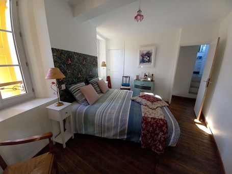 A bedroom with a bed, a nightstand, a chair, a desk, a dresser, a window, and a door.