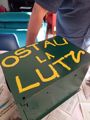 Eine grüne Kiste mit gelbem Text 'OSTALU LA LUTZ' und ein Bein im Hintergrund.