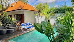 A pool area with lounge chairs, a covered seating area, and lush tropical plants.