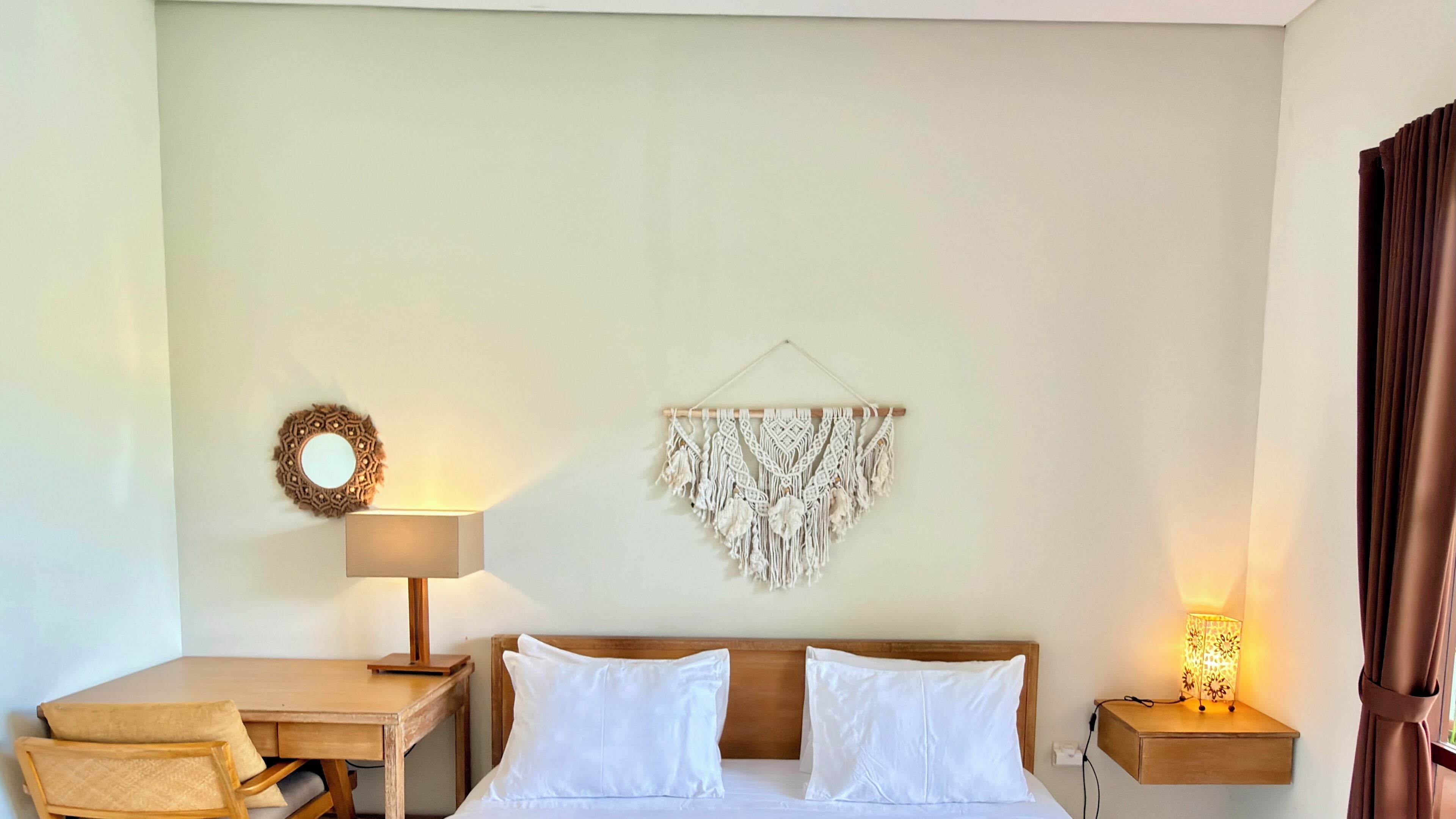 A hotel room with a bed, a desk, a chair, a wall-mounted macrame piece, a wall mirror, and a bedside lamp.