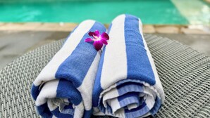 Zwei aufgerollte Handtücher mit blauen und weißen Streifen und einer violetten Blume darauf, auf einem gewobenen Stuhl am Pool platziert.