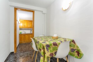 Zona pranzo con tavolo ricoperto da tovaglia a motivi di grappoli d'uva, sedie bianche e un armadietto in legno sullo sfondo.