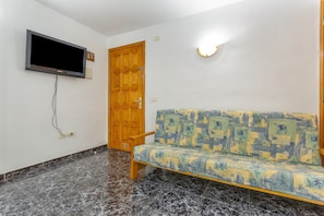 Una stanza con una televisione a parete, una porta in legno, un divano con motivo decorativo e un pavimento piastrellato.