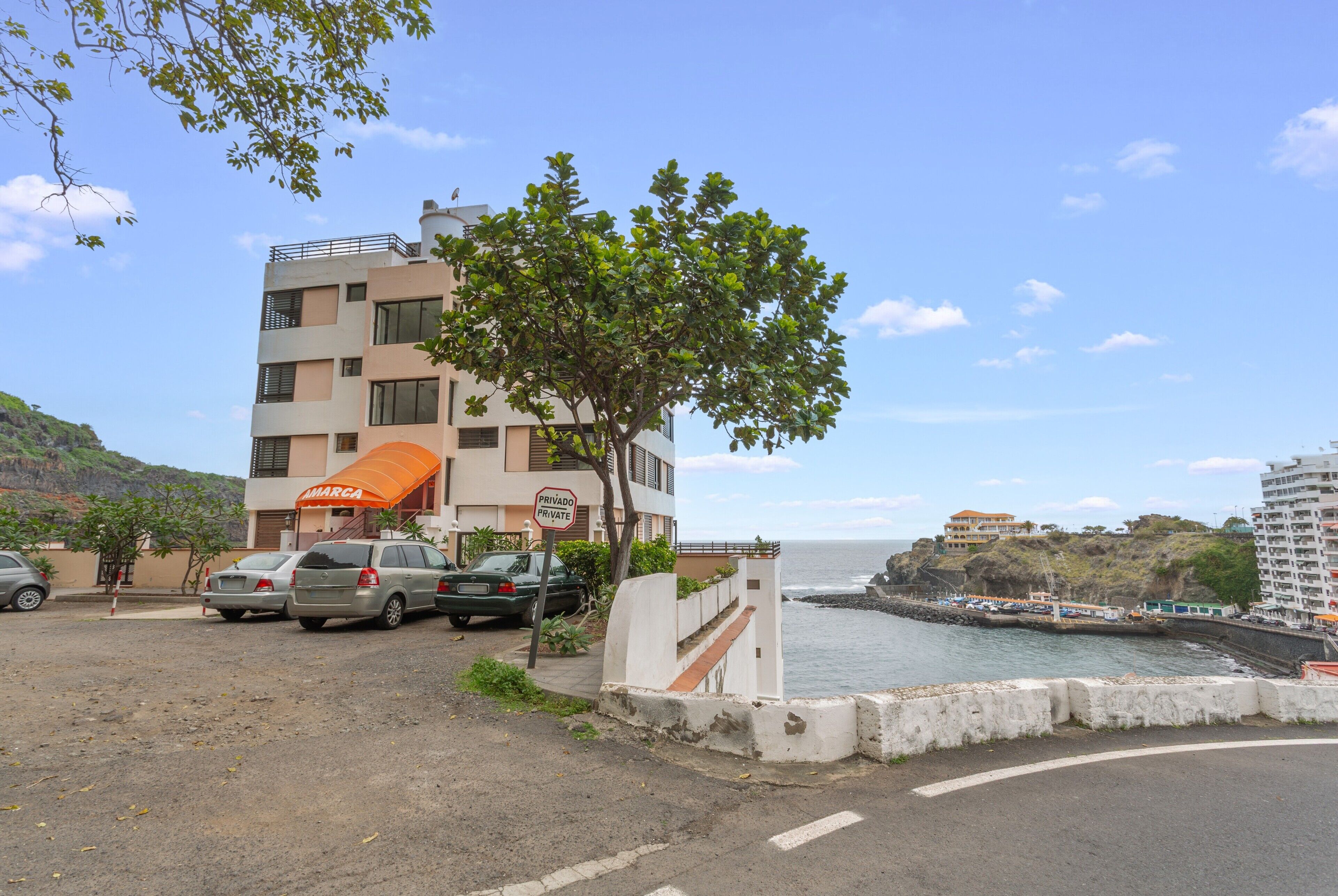 A multi-story building with a sign indicating it is private property, parked cars, a tree, and a view of the ocean.