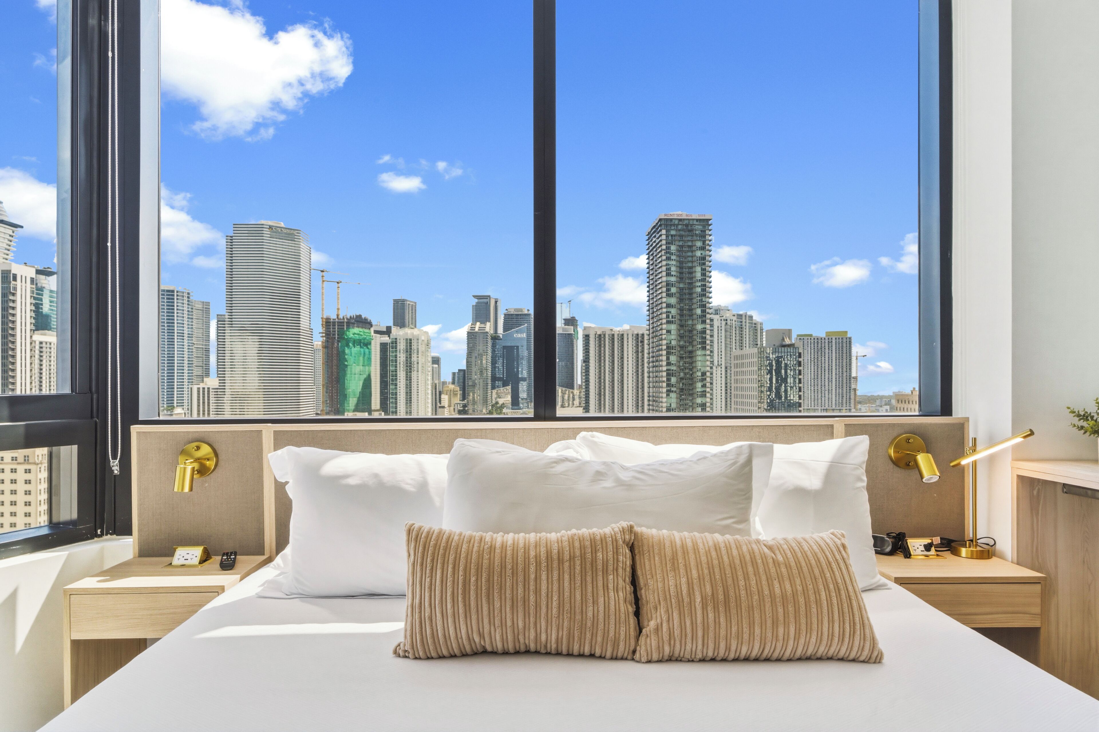 A hotel room with a large window, a bed with white linens, a wooden headboard, a bedside table with a lamp, and a view of a city skyline.