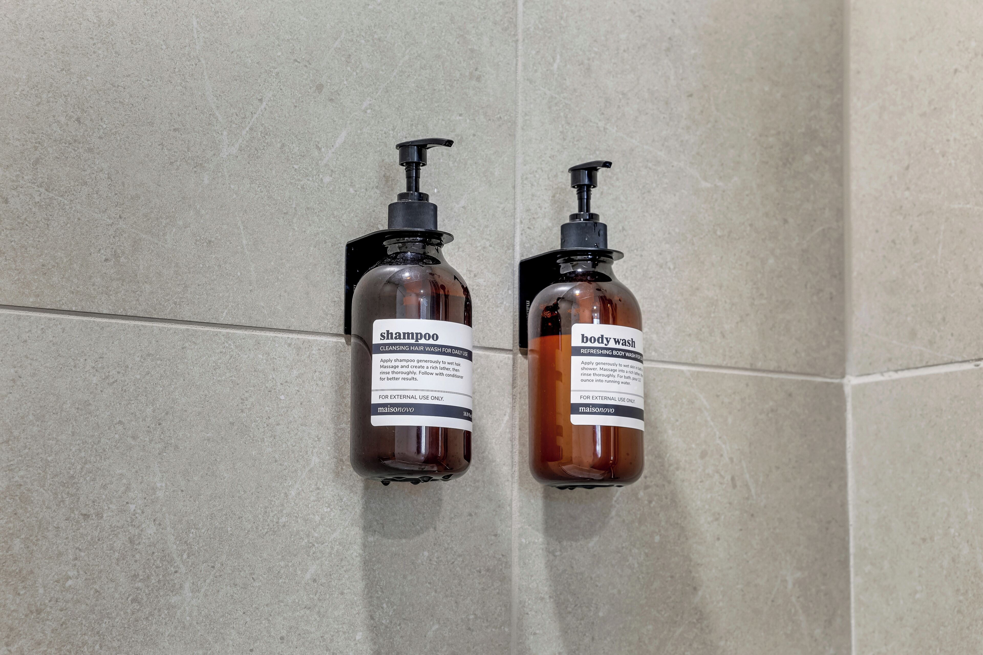 Two bottles of shampoo and body wash on a tiled wall.