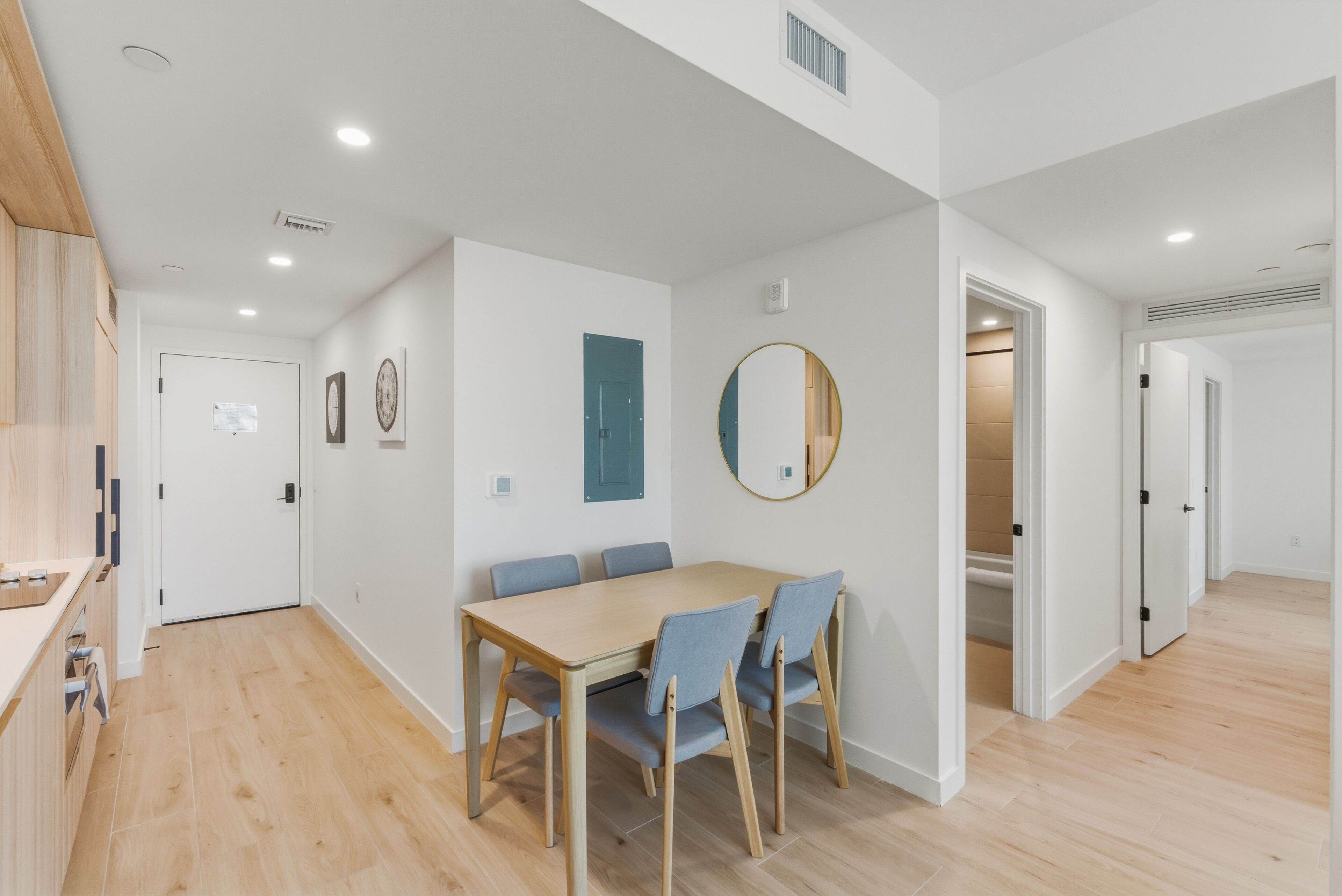 A modern kitchen with wooden cabinets, a dining area with a wooden table and blue chairs, and a hallway with white walls and wooden flooring.