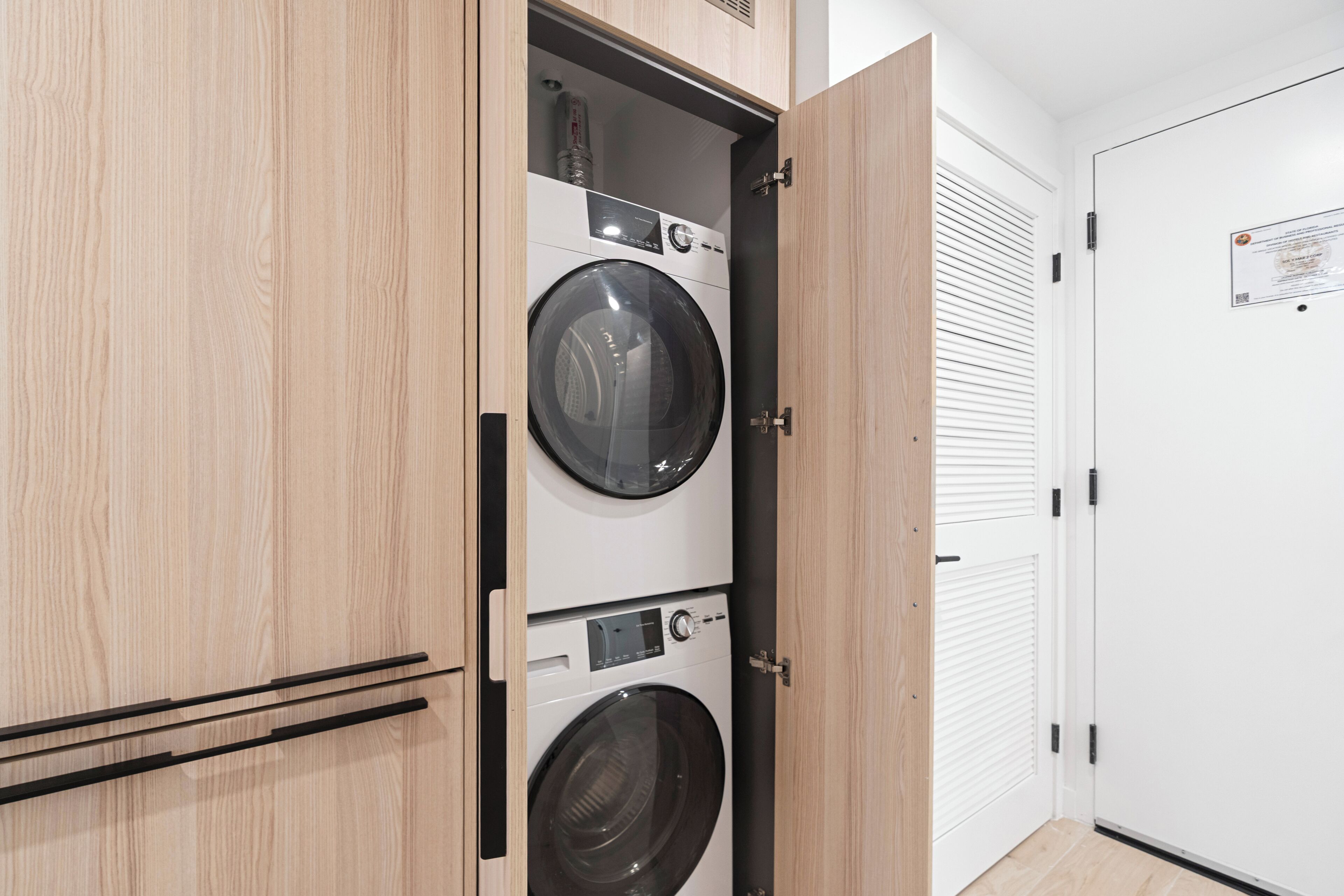 A laundry room with two stacked washing machines, a wooden cabinet, and a white door with a notice on it.