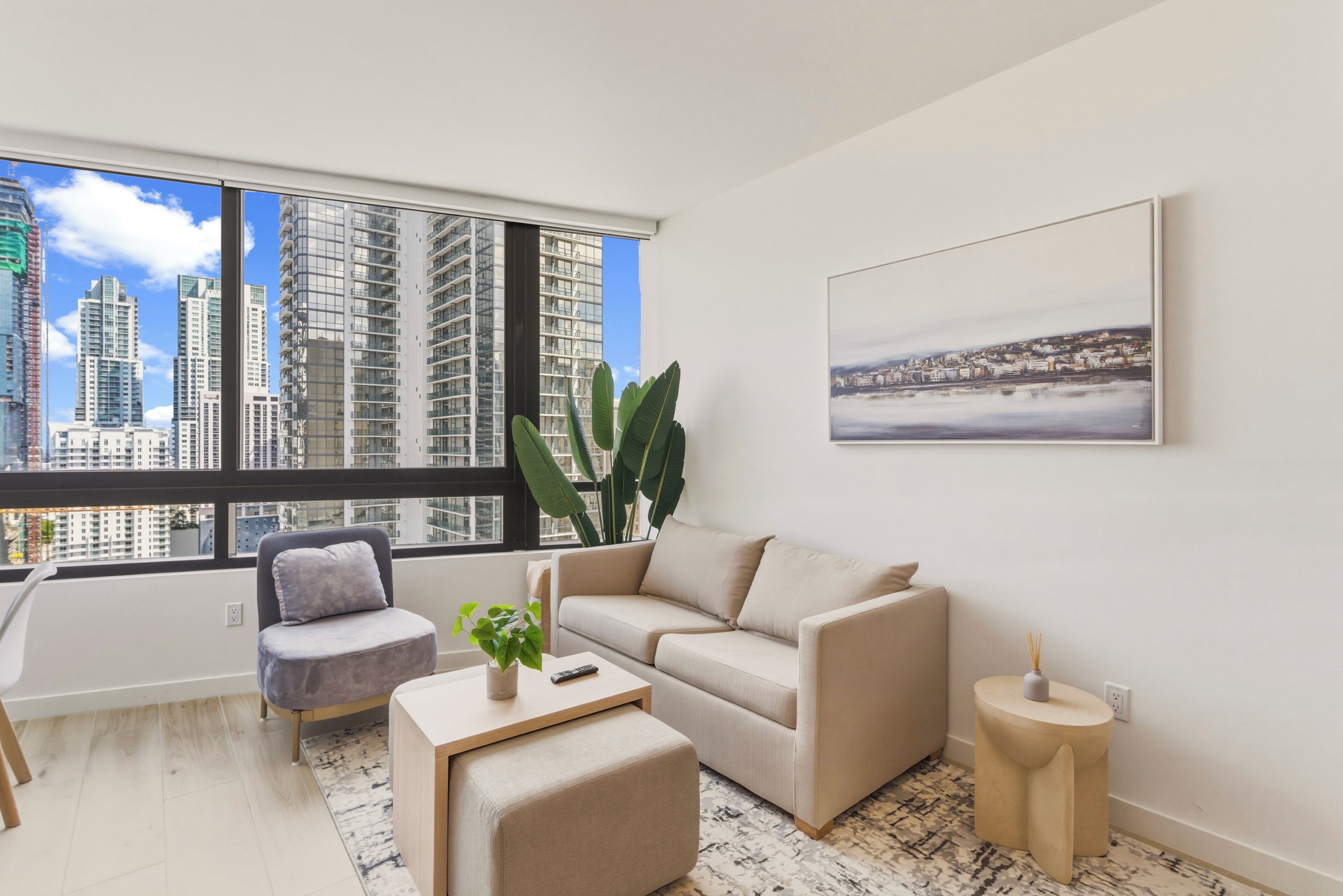 A modern living room with a large window, a sofa, a chair, a small table, and a potted plant.