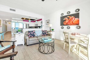 Ein modernes Wohnzimmer mit einem Glastisch, einer Couch mit floralen Kissen, einem Essbereich mit weißem Tisch und Stühlen sowie einer Küche im Hintergrund.