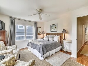 A bedroom with a bed, a ceiling fan, a window with curtains, a bedside table with a lamp, and a chair. - 5BR Beach Home | Steps to Ocean | Sleeps 12! (North Bethany Beach)