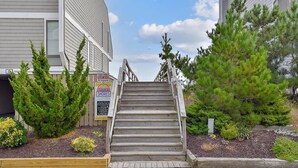 A set of wooden stairs leading to a building entrance, surrounded by greenery and a signboard. - 5BR Beach Home | Steps to Ocean | Sleeps 12! (North Bethany Beach)