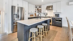 A modern kitchen with white cabinets, a stainless steel refrigerator, a central island with stools, and a built-in oven. - 5BR Beach Home | Steps to Ocean | Sleeps 12! (North Bethany Beach)