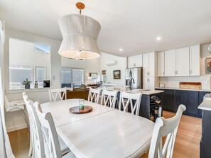 A modern kitchen with white cabinetry, a central island, and a dining area with white chairs. - 5BR Beach Home | Steps to Ocean | Sleeps 12! (North Bethany Beach)