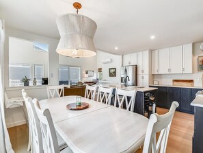 A modern kitchen with white cabinetry, a central island, and a dining area with white chairs. - 5BR Beach Home | Steps to Ocean | Sleeps 12! (North Bethany Beach)