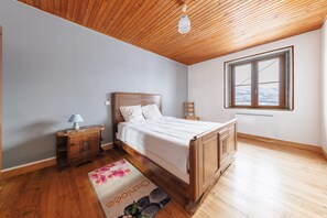 Quarto com teto de madeira, cama com roupa de cama branca, mesa de cabeceira, luminária, janela com vista para montanhas e tapete floral.