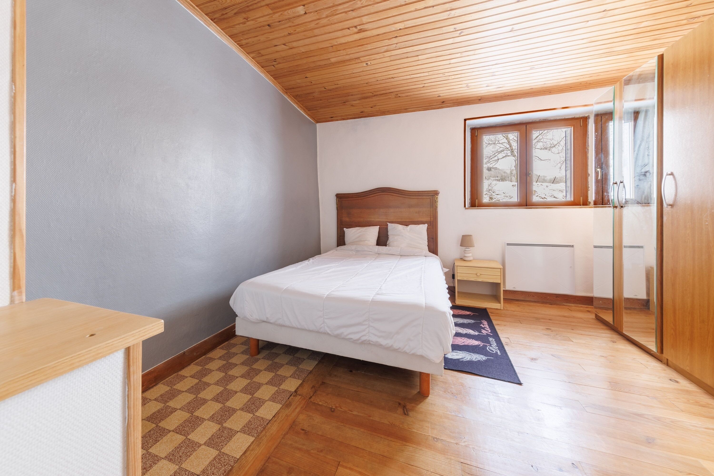 Ein Schlafzimmer mit Holzbalkendecke, einem Bett mit weißer Bettwäsche, einem Schachbrettteppich, einem Nachttisch und einem Fenster mit Blick auf Bäume.