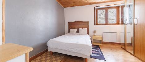 Ein Schlafzimmer mit Holzbalkendecke, einem Bett mit weißer Bettwäsche, einem Schachbrettteppich, einem Nachttisch und einem Fenster mit Blick auf Bäume.