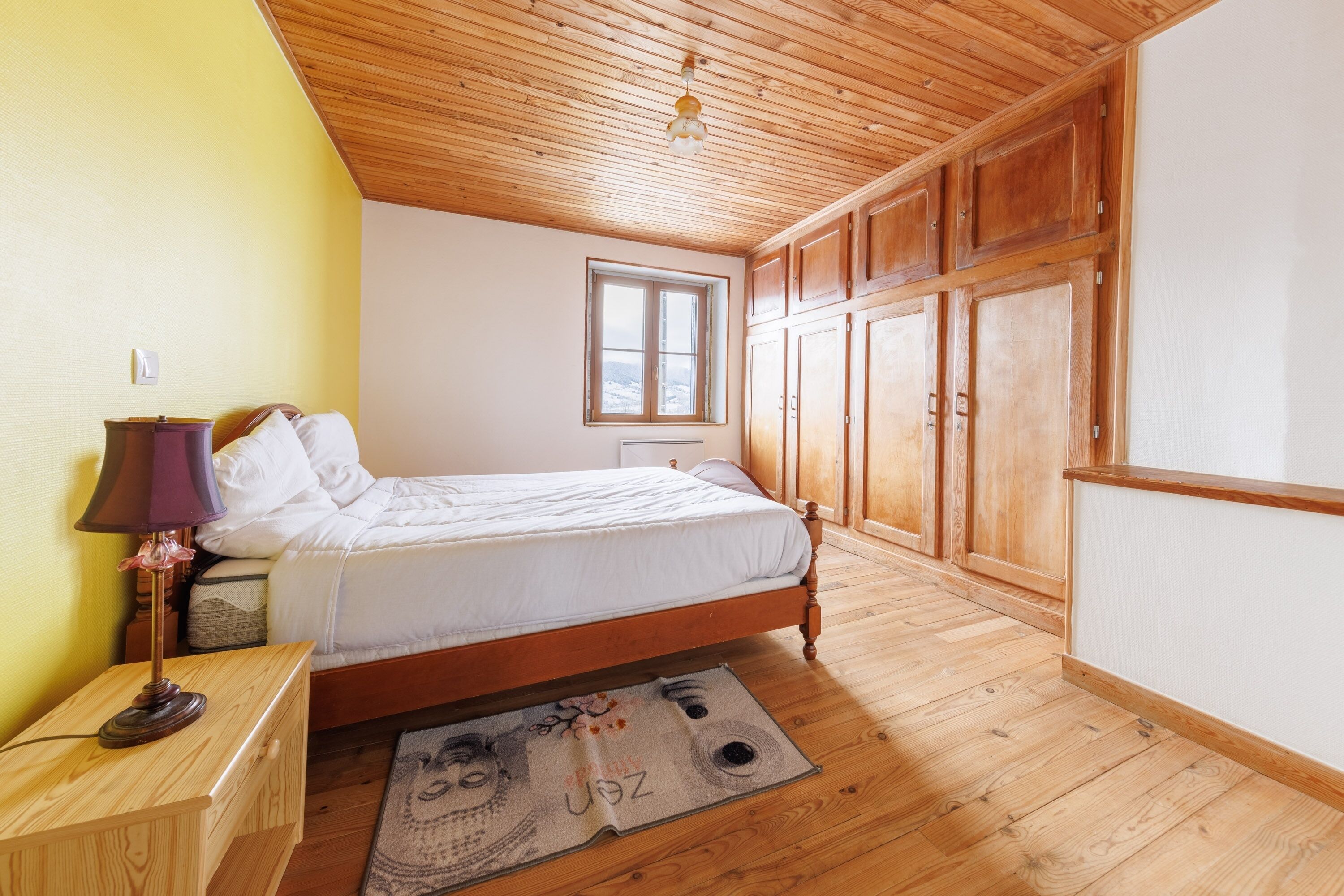 Quarto com teto de madeira, cama, mesa de cabeceira, abajur e janela.