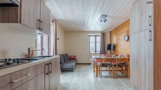 Kompakti keittiö, jossa on puiset kaapit, ruokailutila pöytineen ja tuoleineen, sohva ja televisio.