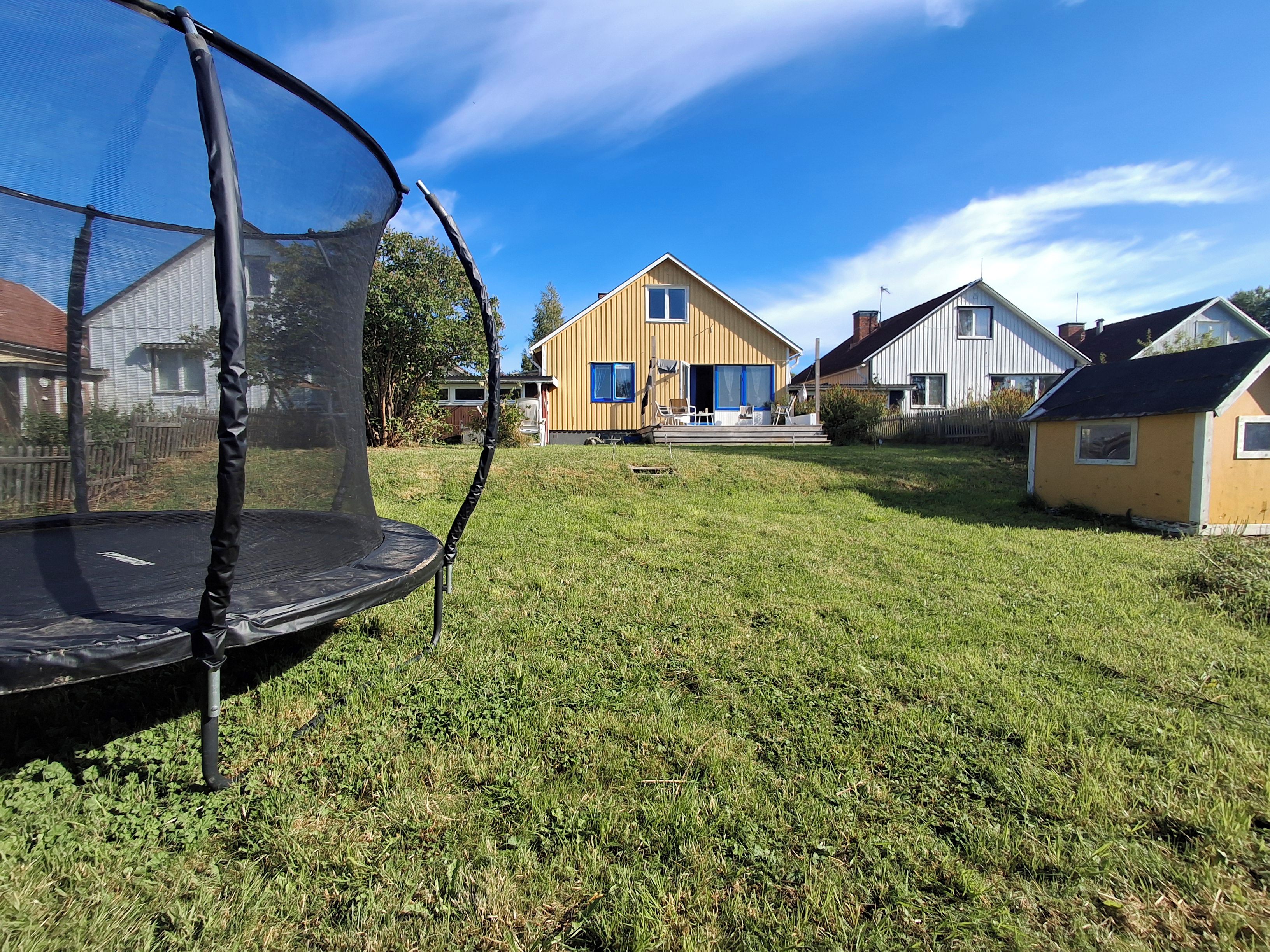 Un trampolín en un jardín con césped y casas al fondo.