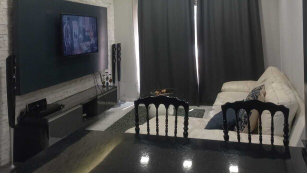 A modern living room with a flat-screen TV mounted on a white brick wall, a black coffee table, a white sofa with patterned cushions, and two red pendant lights. - Lovely 3-bedroom apartment with AC in vibrant Luanda (Luanda)
