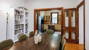 Un coin repas avec une table et des chaises en bois, une bibliothèque et une cuisine visible à travers une porte ouverte.
