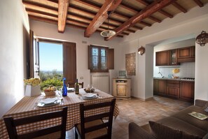 A dining area with a table set for two, a kitchen with wooden cabinets, and a view through an open window. - Agriturismo Fiordaliso (Trevi)