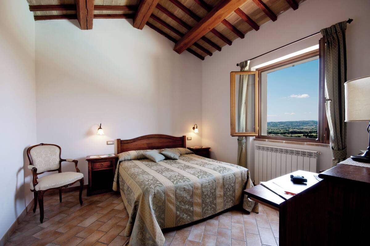 Un dormitorio con techo de madera, una cama con colcha estampada, una silla, un escritorio y una ventana con vistas al campo.