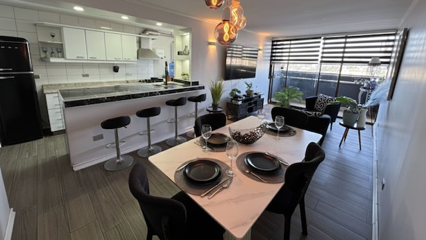 A modern kitchen with white cabinets, a central island with bar stools, and a dining area with a table set for four. - Spacious apartment with panoramic view and connectivity in downtown Santiago (San Miguel)