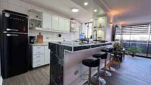 A modern kitchen with a black refrigerator, white cabinets, a marble island with bar stools, and a city view through large windows. - Spacious apartment with panoramic view and connectivity in downtown Santiago (San Miguel)