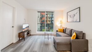 A modern living room with a grey sofa, a small coffee table, a TV on a wooden stand, a large window with a city view, and a framed picture on the wall. - Cozy&Modern New 1 Bedroom Condo (Surrey)