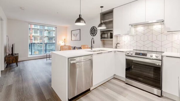 A modern kitchen with white cabinetry, a stainless steel dishwasher, a built-in oven, and a large window providing a view of the cityscape. - Cozy&Modern New 1 Bedroom Condo (Surrey)