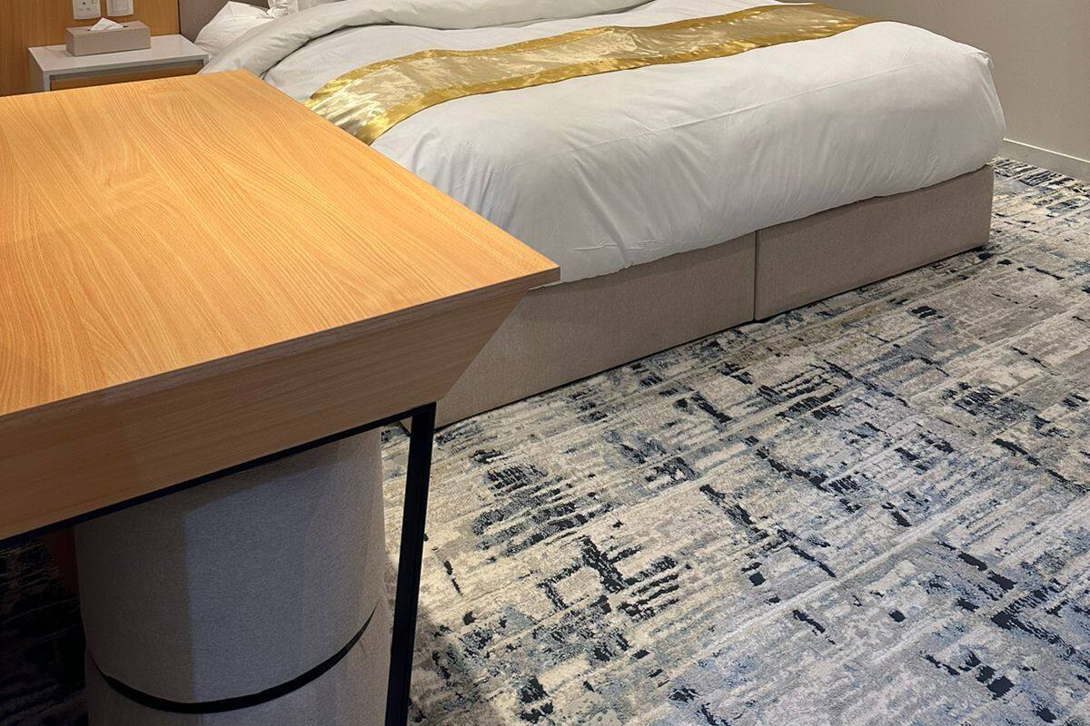 A modern hotel room with a large bed, a wooden nightstand, a desk, and a patterned rug.