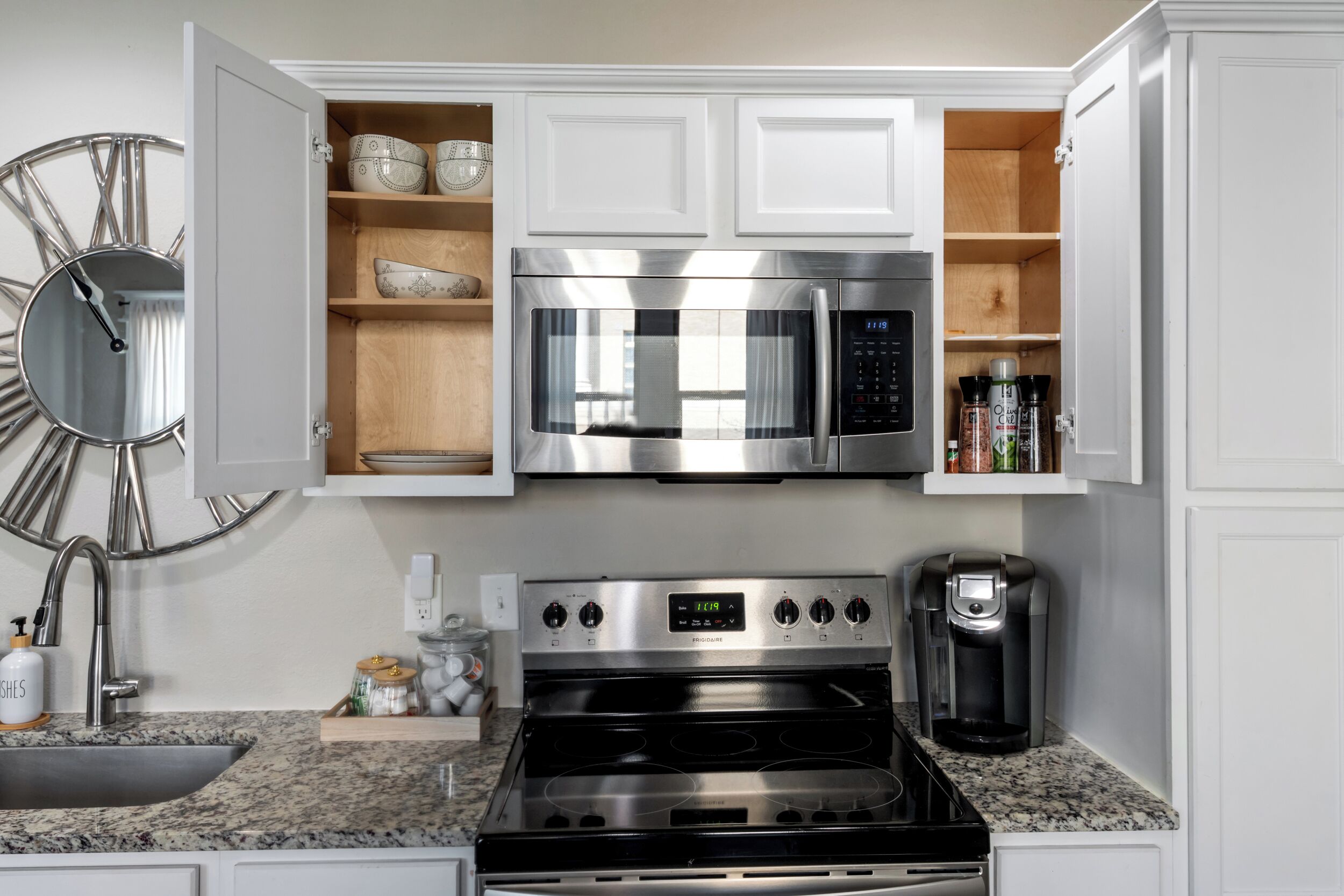 A modern kitchen with white cabinets, a stainless steel microwave, a coffee maker, and a clock on the wall.