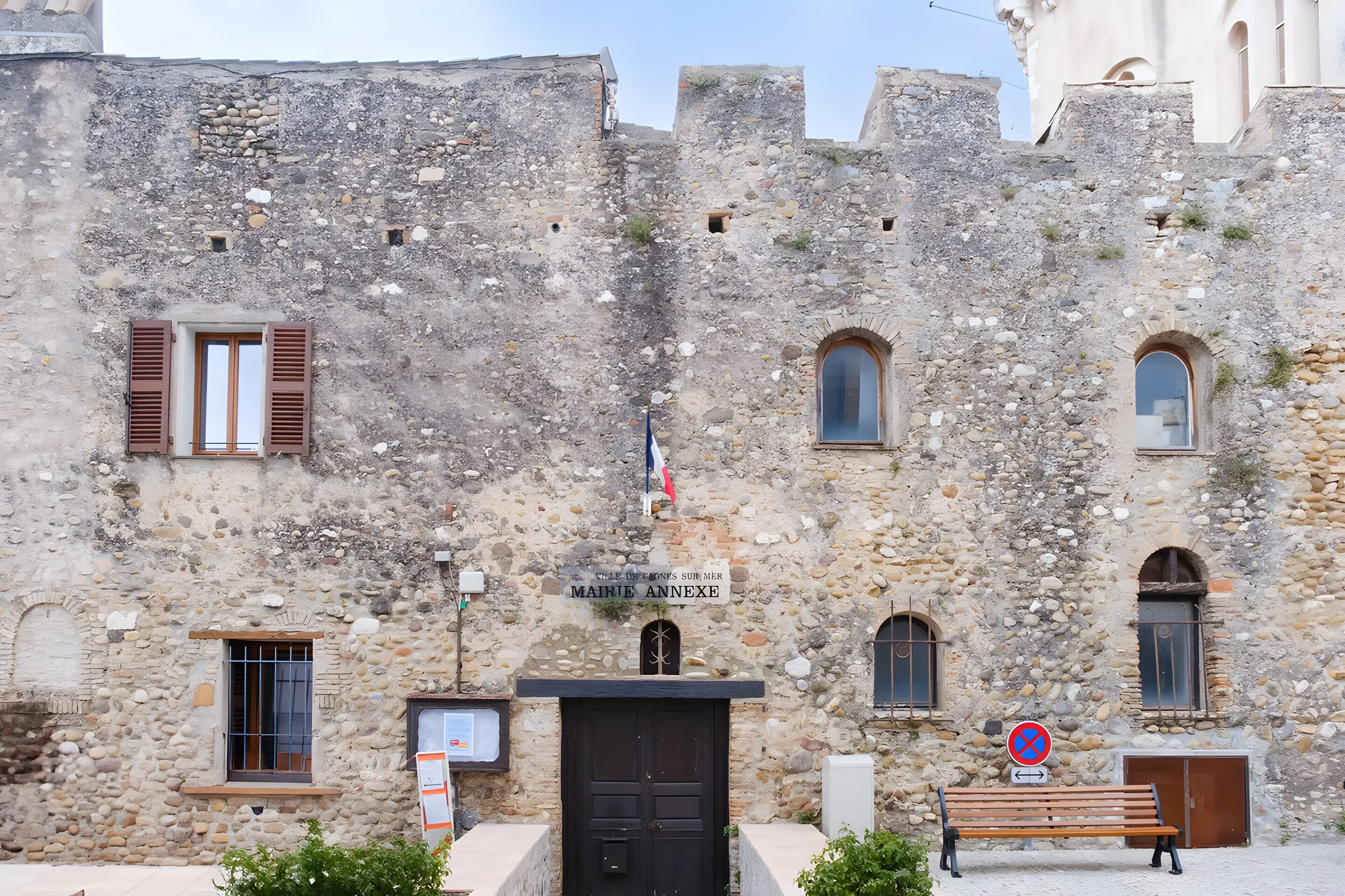 A stone building with a sign reading 'MAIRIE ANNEXE,' a flag, and a bench in front.