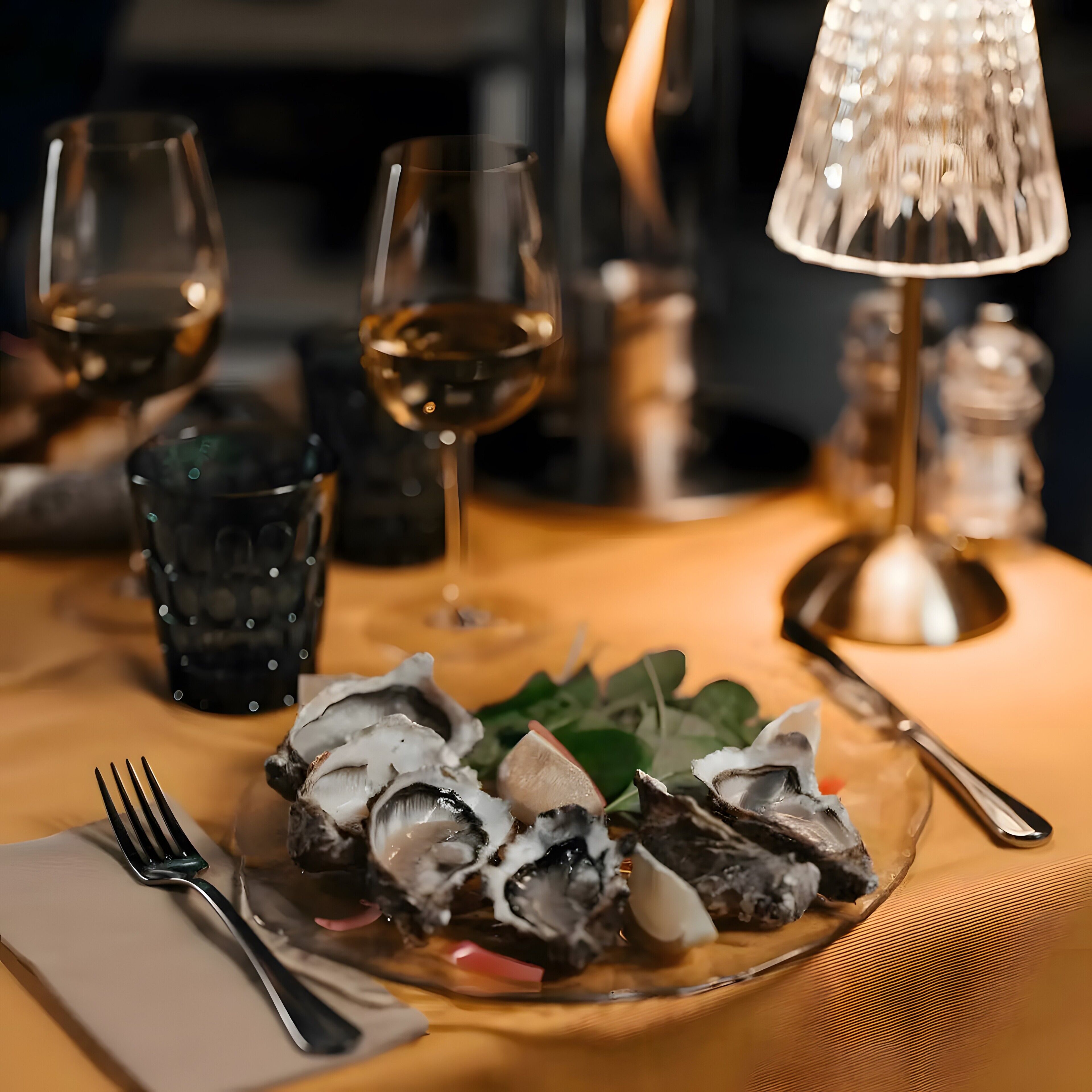 A dining table with a plate of oysters, wine glasses, and a lamp.