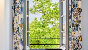 Une pièce avec une fenêtre donnant sur un arbre, des rideaux à motifs floraux et une chaise bleue.