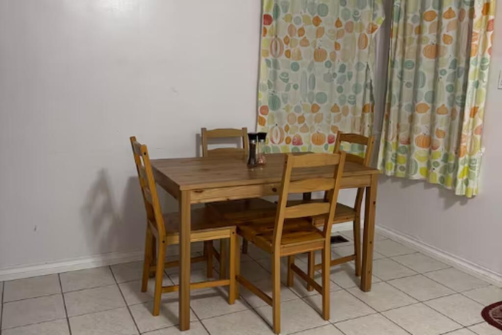 A dining table with four wooden chairs, a small table lamp, and a patterned curtain in the background.